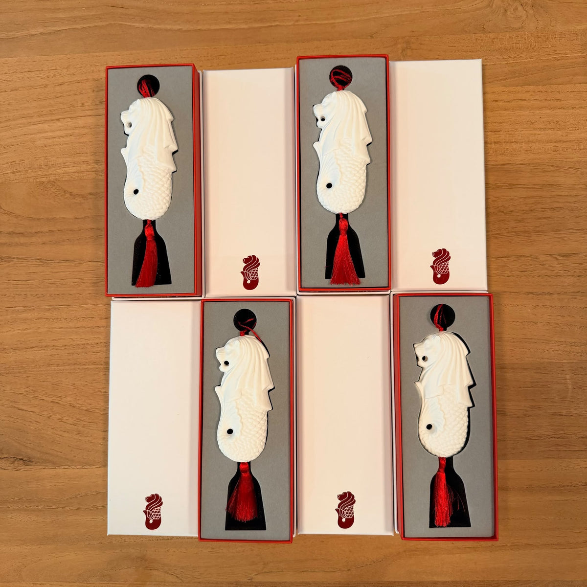 Four White and red presentation gift boxes arranged in a 2x2 grid on a light wood surface. Each box contains a white bone china merlion decoration with a  red tassel base, set against a gray insert with a small red foil merlion emblem near the bottom.