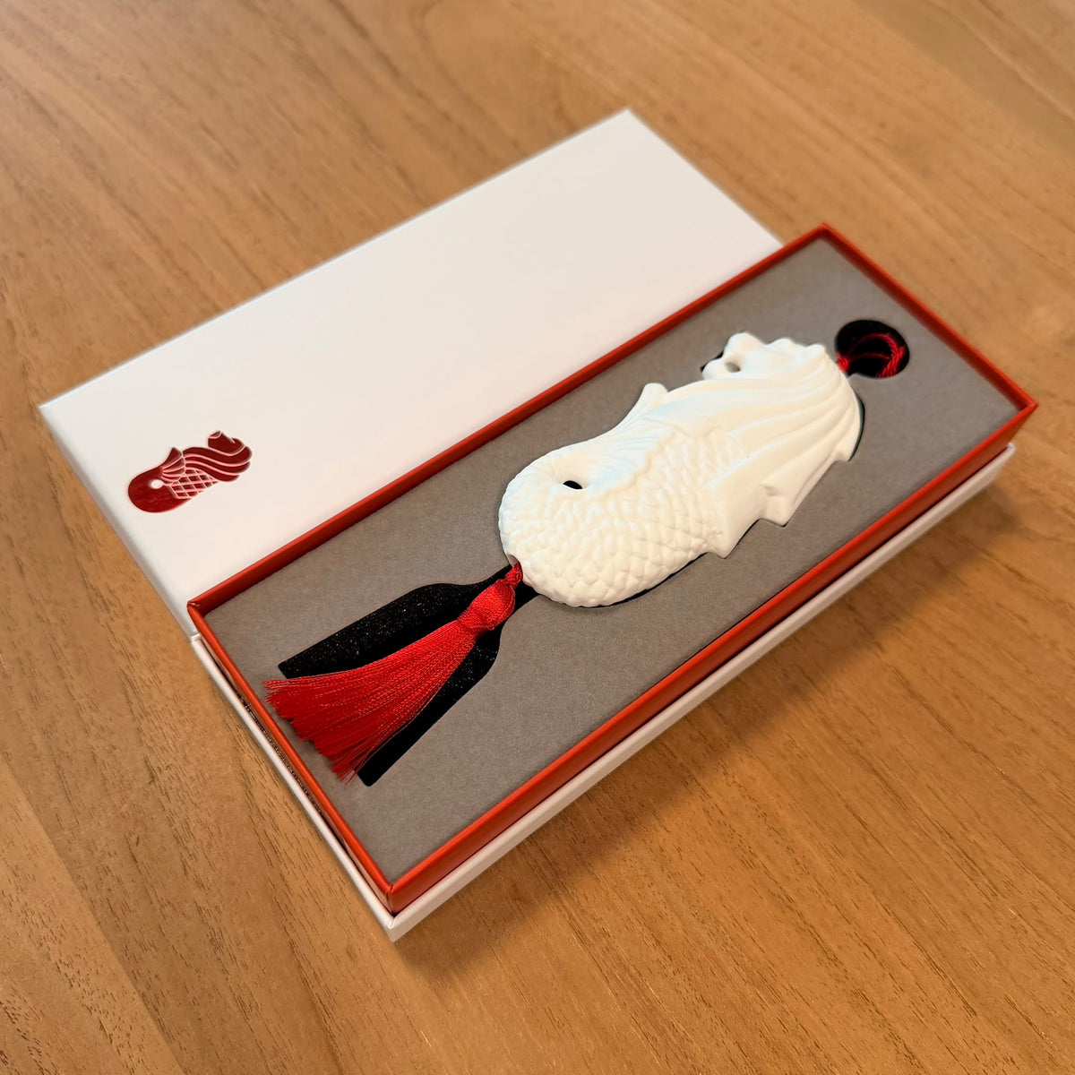 A white bone china merlion hanging ornament with a red tassel, presented in an open white and red box on a wooden table.