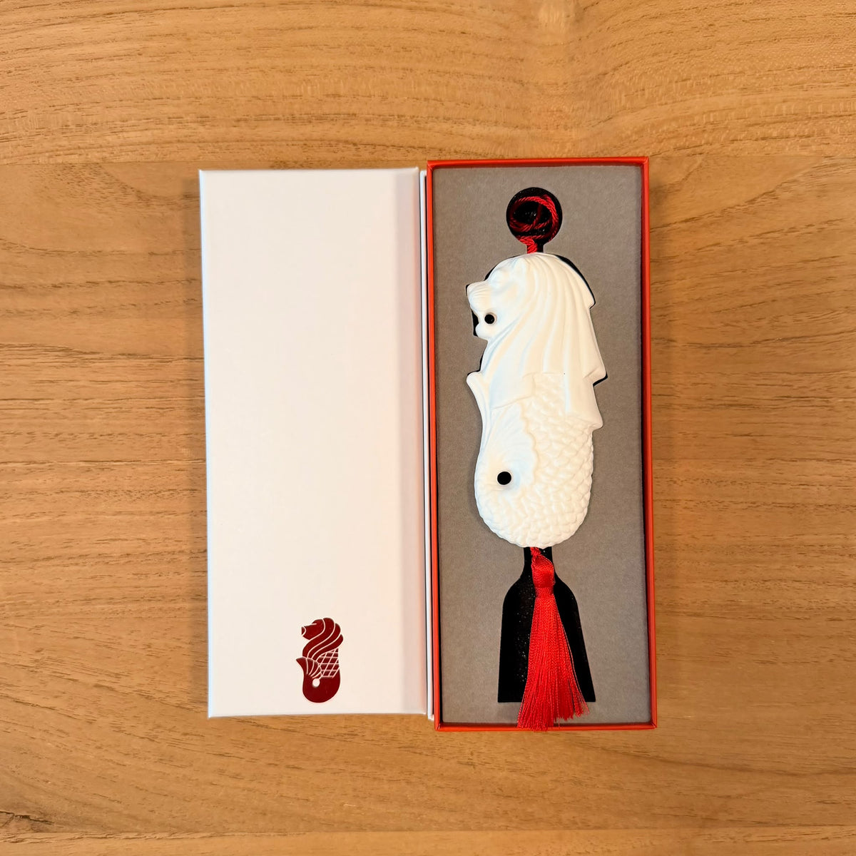 A white bone china merlion ornament with a red tassel, displayed in an open red-bordered presentation box on a light wood surface.